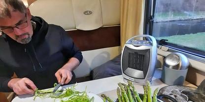 Person mit Brille sitzt an einem Tisch und schneidet gr&uuml;nen Spargel auf einem Schneidebrett; auf dem Tisch stehen ein kleiner Heizl&uuml;fter, eine Schale und Utensilien; Fenster mit Au&szlig;enblick