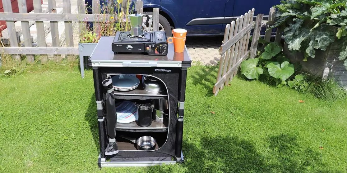 Offener schwarzer Kasten auf Rasen. Auf der Oberseite ein Zweiflamm-Gaskocher und ein orangefarbener Thermobecher. Innen Regalb&ouml;den mit T&ouml;pfen, Pfannen, Tellern und Besteck. Im Hintergrund wei&szlig;er Lattenzaun, Holzzaun, Pflanzen und ein dunkelblaues Au