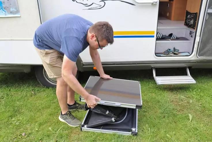 Person tr&auml;gt blaues T-Shirt, kurze Hose und Sneaker und hebt eine klappbare Platte an der Seite eines geparkten Wohnmobils; darunter ein offenes Fach mit Schl&auml;uchen, ge&ouml;ffnete T&uuml;r und Tritt sichtbar