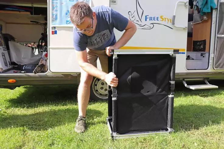 Mann mit Brille und T-Shirt montiert einen schwarzen, zusammenklappbaren Kasten vor einem wei&szlig;en Wohnwagen auf Gras