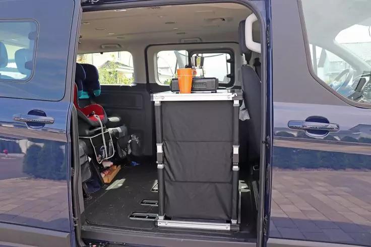 Offene Seitenschiebet&uuml;r eines dunkelblauen Vans; Blick in den Innenraum mit schwarzem rechteckigem Stoffpaneel auf Schienen, orangefarbener Becher auf einer wei&szlig;en Ablage, links ein Sitz mit rotschwarzem Kindersitz, Pflaster drau&szlig;en