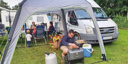 Grau-wei&szlig;er Campingbus neben einem wei&szlig;en Vorzelt; mehrere Personen sitzen an einem Tisch unter dem Zelt; ein Mann hockt vor einer K&uuml;hlbox und einem tragbaren Gaskocher; eine Propangasflasche steht auf dem Rasen