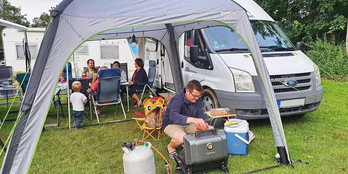 Grau-wei&szlig;er Campingbus neben einem wei&szlig;en Vorzelt; mehrere Personen sitzen an einem Tisch unter dem Zelt; ein Mann hockt vor einer K&uuml;hlbox und einem tragbaren Gaskocher; eine Propangasflasche steht auf dem Rasen