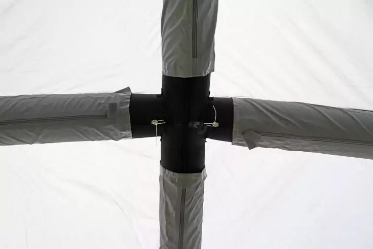 Schwarzes Kreuzverbindungsstück, an dem vier graue Stangen mit Stoffhüllen befestigt sind; zwei kleine Metallklammern sichtbar; heller Hintergrund