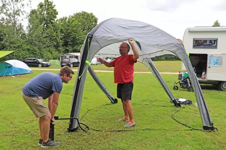 Zwei Männer unter grauem aufblasbarem Pavillon auf einer Wiese; einer in rotem T‑Shirt und schwarzen Shorts hält eine Stange, der andere in blauem T‑Shirt und beigen Shorts beugt sich zum Boden; Wohnwagen rechts, Zelt und Autos links, Bäume im Hinter