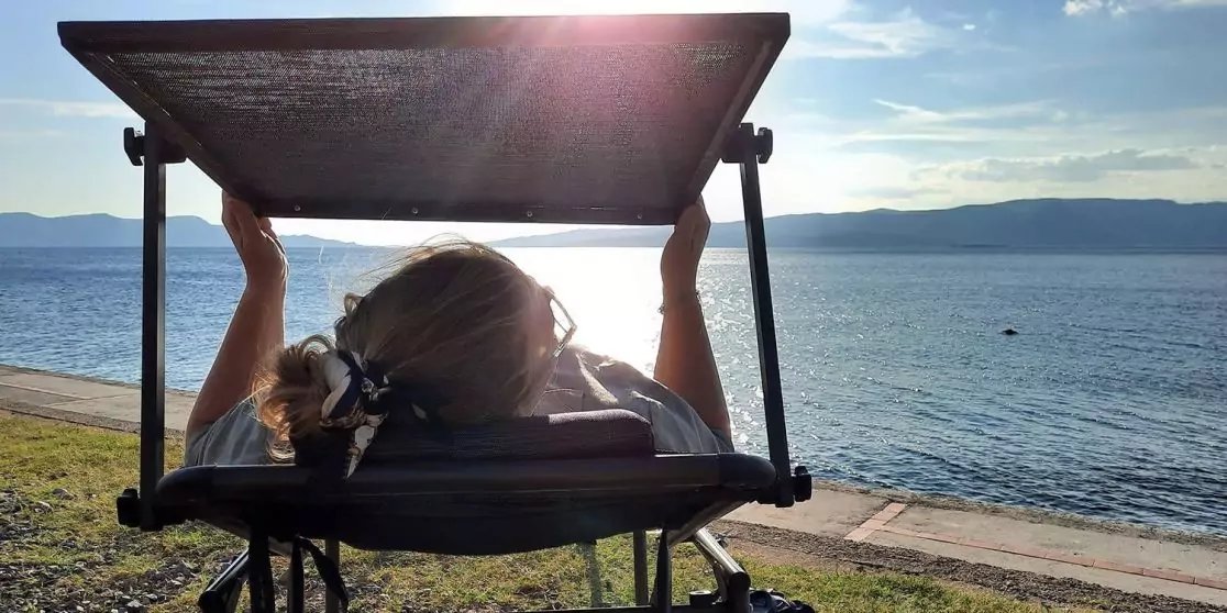 R&uuml;ckansicht einer Person in einem Liegestuhl mit Sonnendach, blickt aufs Meer; Strand, entfernte H&uuml;gel und sonniger Himmel sichtbar