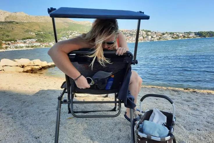 Person mit blondem Haar beugt sich nach vorn in einer Strandliege, neben ihr ein Kinderwagen, im Hintergrund Meer, Küste und Häuser