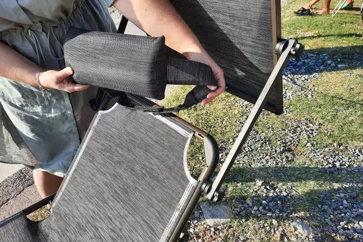 Hände halten ein dunkles, gepolstertes Kopfkissen über der Lehne eines grauen Liegestuhls mit Aluminiumrahmen; zweiter Liegestuhl daneben, Kies und Rasen im Hintergrund