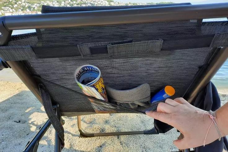 Rückseite eines Strandstuhls mit Stofftasche; darin eine zusammengerollte Zeitschrift und eine Flasche mit orangefarbenem Deckel. Eine Hand mit Armband greift in die Tasche. Sand und Wasser im Hintergrund