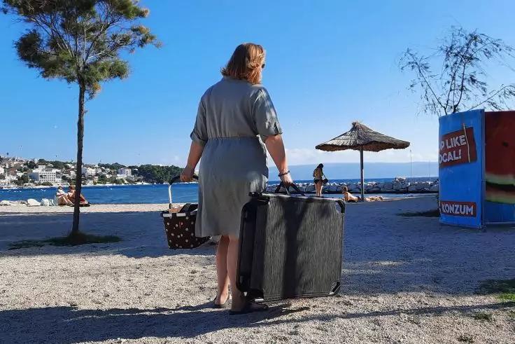 Person von hinten in hellgrauem Kleid am Strand mit großem Rollkoffer und Korb; Meer, Sonnenschirm, Kiosk, Häuser am gegenüberliegenden Ufer und blauer Himmel