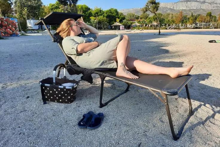 Eine Person liegt auf einer Liege am sandigen Ufer und hält eine Hand vor die Stirn; daneben eine gepunktete Tasche und Sandalen, im Hintergrund Bäume und Berge