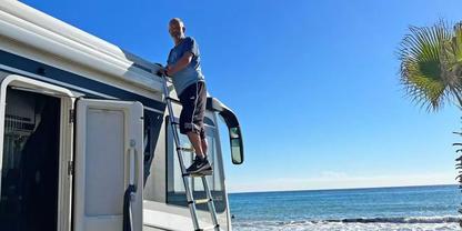 Person steht auf einer Leiter, die an einem Wohnmobil befestigt ist; Meer und blauer Himmel im Hintergrund, Palme am rechten Bildrand
