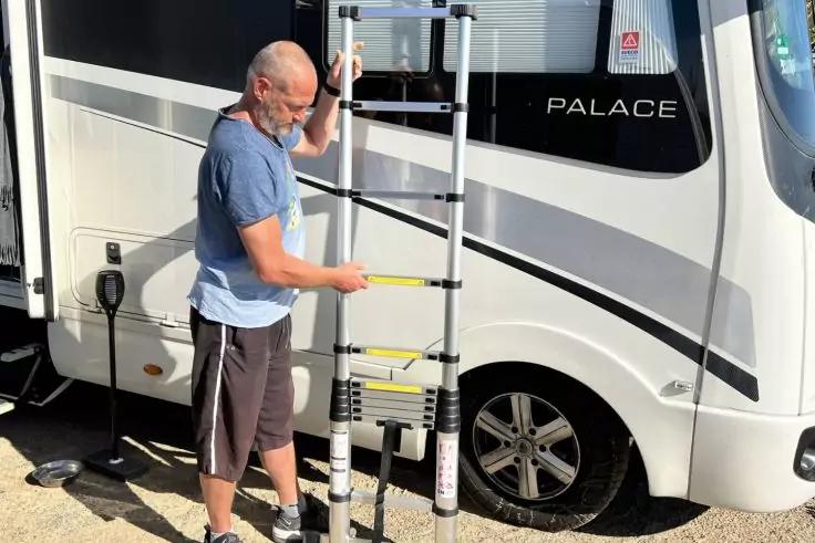 Ein Mann h&auml;lt eine Teleskopleiter neben einem wei&szlig;en Wohnmobil mit dem Schriftzug "PALACE"; Rad und ein Stromkabel sind sichtbar
