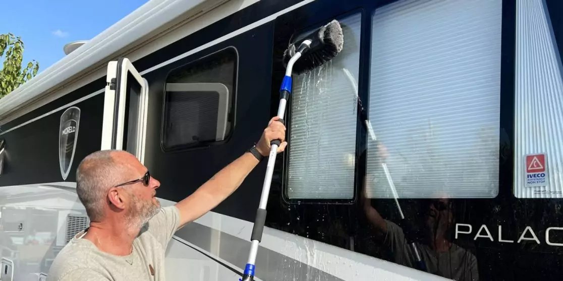 Mann mit kurzem Haar und Sonnenbrille h&auml;lt eine Teleskopb&uuml;rste und w&auml;scht die Au&szlig;enscheiben eines Wohnmobils