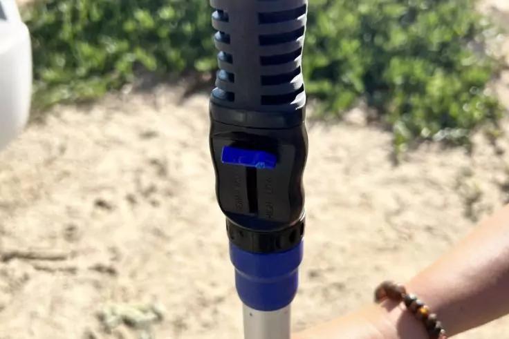 Nahaufnahme eines schwarz-blauen Kunststoffverbinders an einer silbernen Stange, im Hintergrund Sand und Gr&uuml;n, rechts unten ein Arm mit Perlenarmband