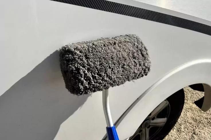 Grauer Farbroller an der Seitenwand eines wei&szlig;en Fahrzeugs nahe dem Radhaus, Reifen und Felge teilweise sichtbar, Kiesboden und Schatten