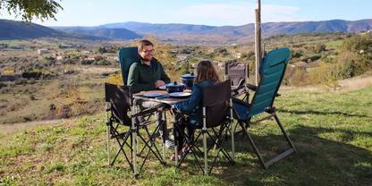 Zwei Personen sitzen an einem Klapptisch zwischen Campingst&uuml;hlen auf einer Wiese, Teller und Tassen auf dem Tisch, im Hintergrund Tal mit H&auml;usern und Bergen