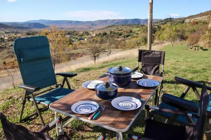 Ein Tisch auf einer Wiese mit vier St&uuml;hlen, zwei T&ouml;pfen und vier Tellern; im Hintergrund H&uuml;gel und Himmel