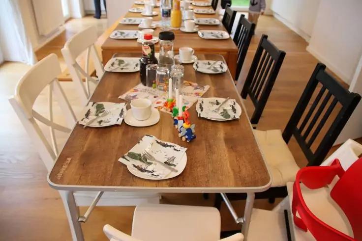 Langer Holztisch mit Tellern, Servietten, Tassen, Salz- und Pfefferstreuern und kleinem Spielzeug in der Mitte; verschiedene St&uuml;hle, darunter ein roter Hochstuhl