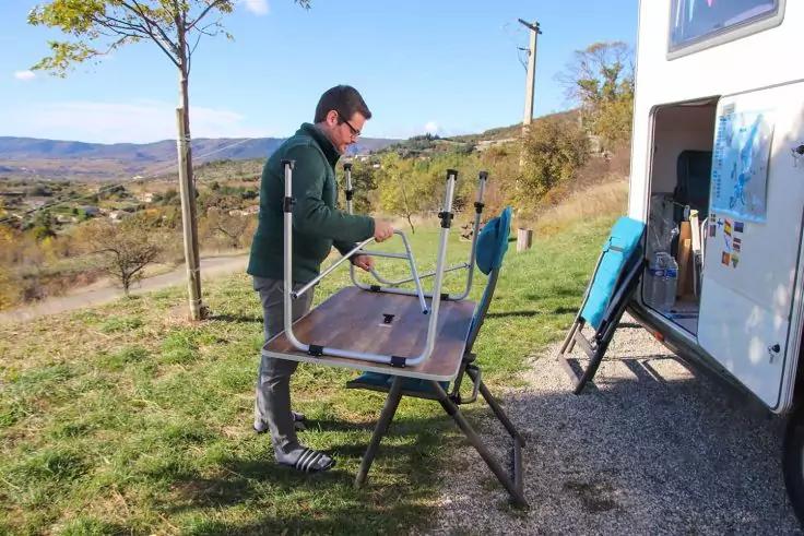 Ein Mann in Jacke und Sonnenbrille steht neben einem offenen Wohnmobil und h&auml;lt eine Tischplatte auf einem klappbaren Gestell; H&uuml;gel und B&auml;ume im Hintergrund