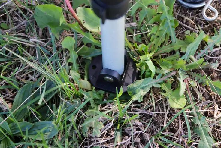 Hellgrauer Stab mit runder schwarzer Fu&szlig;platte auf gr&uuml;nen Bl&auml;ttern und trockenem Gras; rotes Kabel am oberen Bildrand