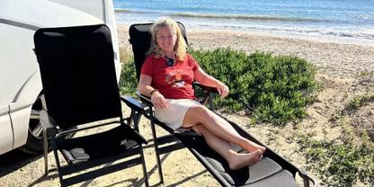 Person mit blondem Haar sitzt auf einem Liegestuhl vor einem wei&szlig;en Wohnmobil am sandigen Strand, Meer und niedrige Vegetation im Hintergrund