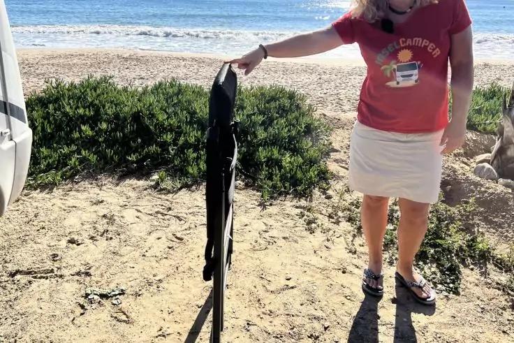 Person von den Schultern abw&auml;rts auf sandigem Strand, h&auml;lt ein zusammengeklapptes schwarzes l&auml;ngliches Objekt an einem St&auml;nder; im Hintergrund Strandvegetation und Meer