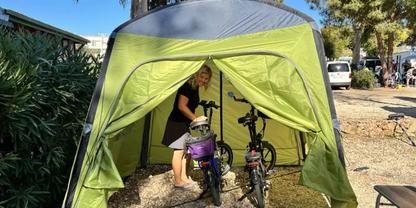 Person steht in offenem gr&uuml;nen Zelt und h&auml;lt ein violettes Kinderfahrrad; daneben ein weiteres Fahrrad; Kiesboden; B&auml;ume, Autos und Geb&auml;ude im Hintergrund