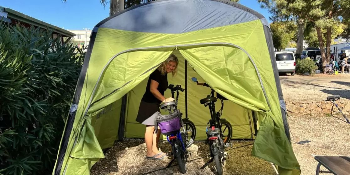 Person steht in offenem gr&uuml;nen Zelt und h&auml;lt ein violettes Kinderfahrrad; daneben ein weiteres Fahrrad; Kiesboden; B&auml;ume, Autos und Geb&auml;ude im Hintergrund