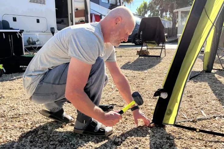 Person kniet auf kiesigem Boden und schl&auml;gt mit Hammer einen Zelthering neben einem gr&uuml;nen Zeltteil ein; Wohnwagen im Hintergrund