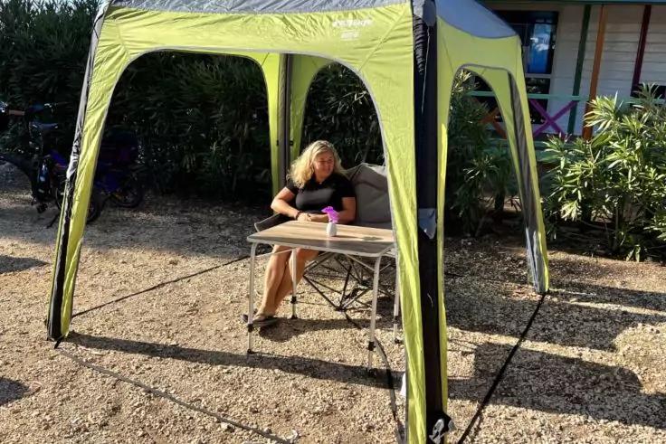 Person mit blonden Haaren sitzt an einem Campingtisch unter gr&uuml;nem Pavillon und h&auml;lt einen violetten Becher; Kiesboden, B&uuml;sche und Geb&auml;ude im Hintergrund, Fahrradrad seitlich sichtbar