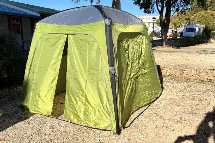 Gr&uuml;n-graues Pop-up-Zelt mit ge&ouml;ffnetem Eingangsflappen auf kiesigem Untergrund, Geb&auml;ude links, Wohnmobil und B&auml;ume im Hintergrund, Schatten einer Person unten rechts