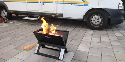 Kleine schwarze Feuerstelle mit brennenden Flammen auf gepflastertem Boden vor einem wei&szlig;en Wohnmobil; Rad, Trittstufe und ein Holzst&uuml;ck sichtbar