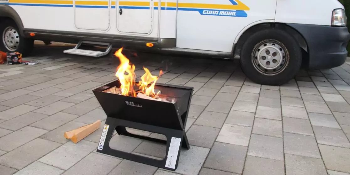 Kleine schwarze Feuerstelle mit brennenden Flammen auf gepflastertem Boden vor einem wei&szlig;en Wohnmobil; Rad, Trittstufe und ein Holzst&uuml;ck sichtbar