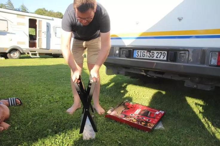 Eine Person beugt sich barfu&szlig; &uuml;ber ein zusammengeklapptes X-f&ouml;rmiges Gestell auf Gras; offene rote Werkzeugkiste und Wohnmobile im Hintergrund