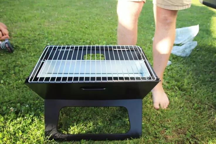 Kleiner schwarzer tragbarer Grill mit Metallrost auf gr&uuml;nem Rasen, dahinter Beine in hellen Shorts und eine Plastikt&uuml;te auf dem Boden