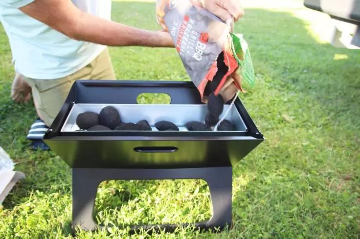 Person gie&szlig;t runde Kohlebriketts aus einer T&uuml;te in einen schwarzen tragbaren Grill auf Rasen; Torso und Arme, offene T&uuml;te, Briketts und Gras sichtbar