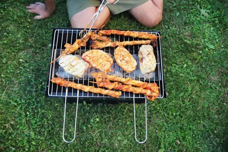 Kleiner tragbarer Grill auf Rasen mit mehreren Brotscheiben und langen Fleischspie&szlig;en auf dem Rost; Oberschenkel und H&auml;nde einer knienden Person oberhalb des Grills sichtbar