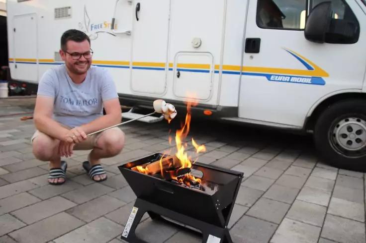 Eine Person mit Brille hockt auf gepflastertem Boden neben einem tragbaren Feuerkorb und h&auml;lt zwei Spie&szlig;e mit Marshmallows &uuml;ber offene Flammen; im Hintergrund ein wei&szlig;es Wohnmobil mit gelben und blauen Streifen