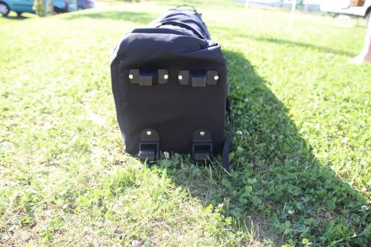 Dunkles rollbares Gep&auml;ckst&uuml;ck liegt auf gr&uuml;nem Rasen, R&uuml;ckseite mit zwei Rollen und zwei kleinen F&uuml;&szlig;en sichtbar, Sonnenlicht und Schatten, unscharfer Hintergrund