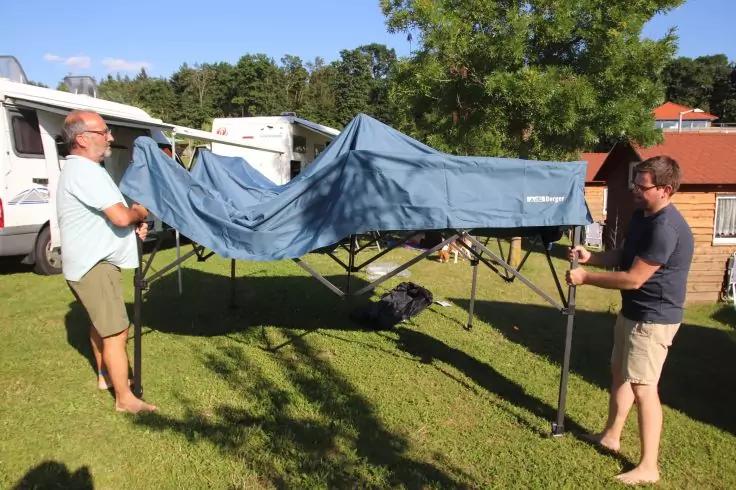 Zwei M&auml;nner halten auf einer Wiese ein teilweise zusammengeklapptes blaues Zeltgestell; im Hintergrund Wohnmobile, B&auml;ume und ein rotes Geb&auml;ude