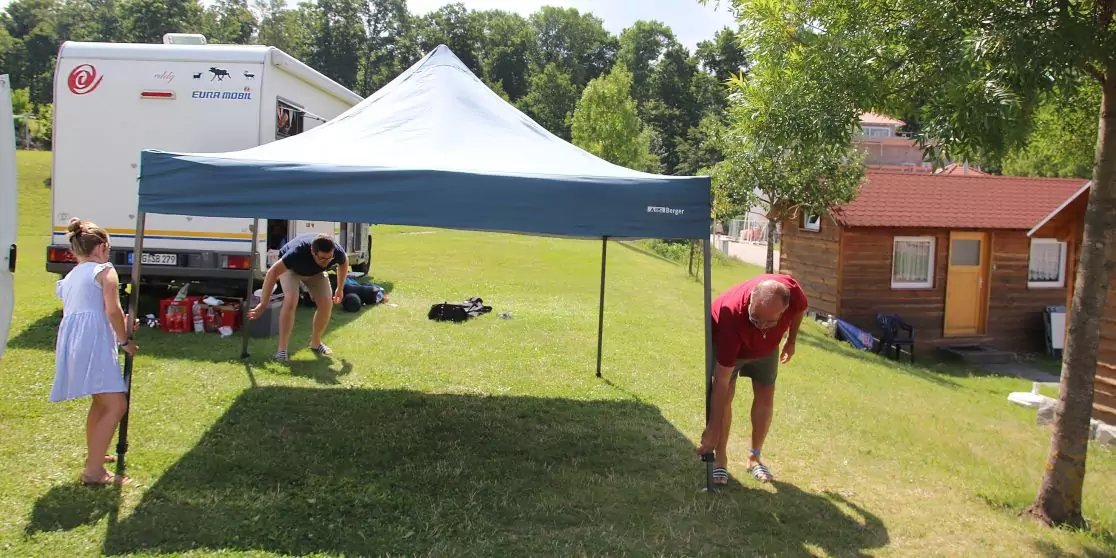 Blaues Faltzelt auf Rasen; links stehende Person in hellblauem Kleid; rechts b&uuml;ckende Person in rotem T‑Shirt an Zeltbein; unter dem Zelt hockende Person; links im Hintergrund wei&szlig;er Wohnwagen; rechts kleines Holzhaus; B&auml;ume im Hintergrund