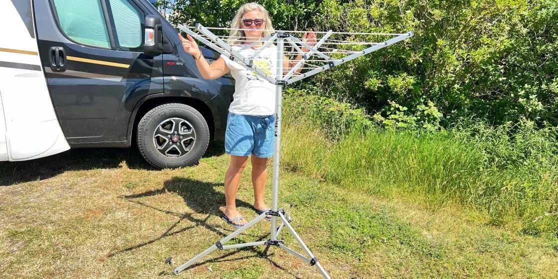 Eine Frau mit Sonnenbrille, wei&szlig;em T-Shirt und blauen Shorts steht auf Gras neben einem ausgeklappten, metallenen W&auml;schetrockner auf Dreibein; im Hintergrund ein Wohnmobil und B&auml;ume