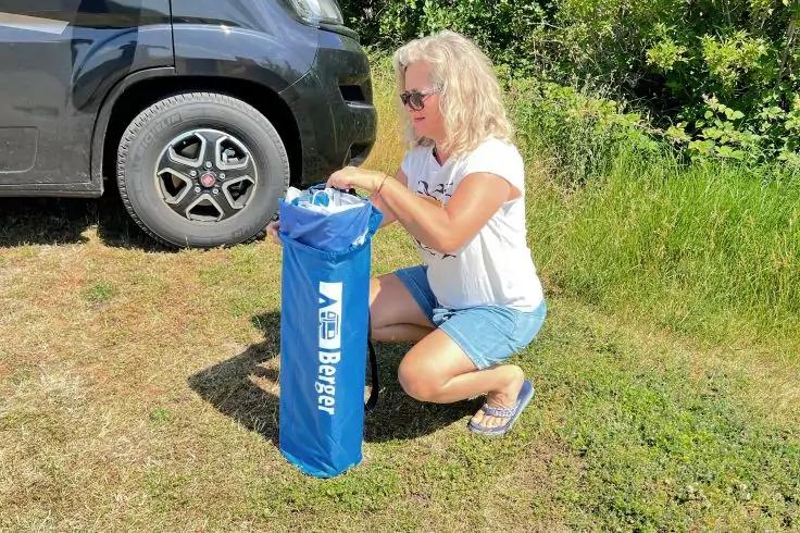 Person kniet auf Gras neben einem grauen Auto und öffnet eine blaue Tasche mit weißem Schriftzug