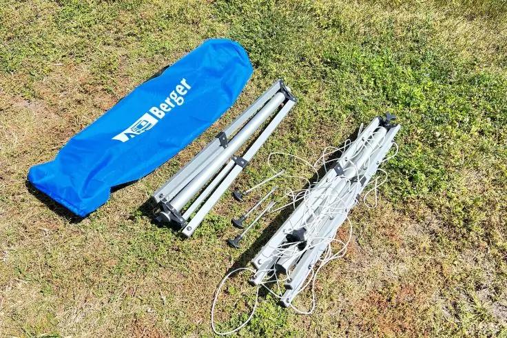 Blaue Tragetasche mit weißer Aufschrift 'Berger', zusammengelegte silberne Metallstangen und zusammengefalteter weißer Metallrahmen mit Seilen, auf Gras