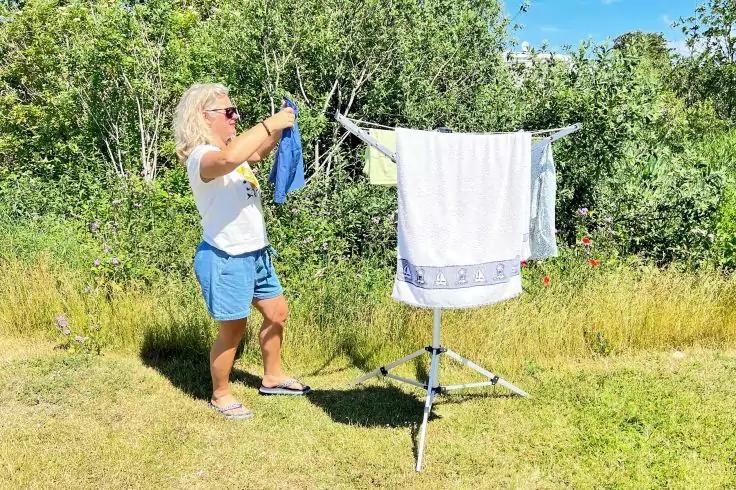 Person hängt ein blaues Kleidungsstück an einem freistehenden Wäscheständer auf einer Wiese; im Hintergrund Büsche und blauer Himmel