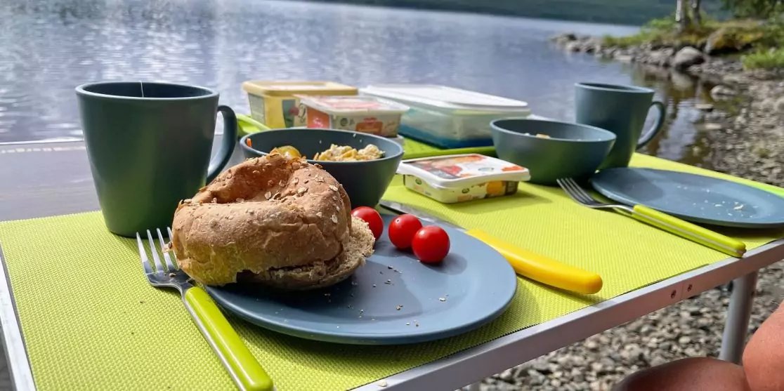Kleiner Tisch mit gr&uuml;nen Platzsets, blauem Teller mit Br&ouml;tchen und zwei kleinen roten Tomaten, Besteck mit gelben Griffen, dunkle Tassen, kleine Sch&uuml;ssel mit hellem Inhalt, rechteckige Aufstrichbeh&auml;lter, See und felsiger Uferabschnitt im Hintergrund
