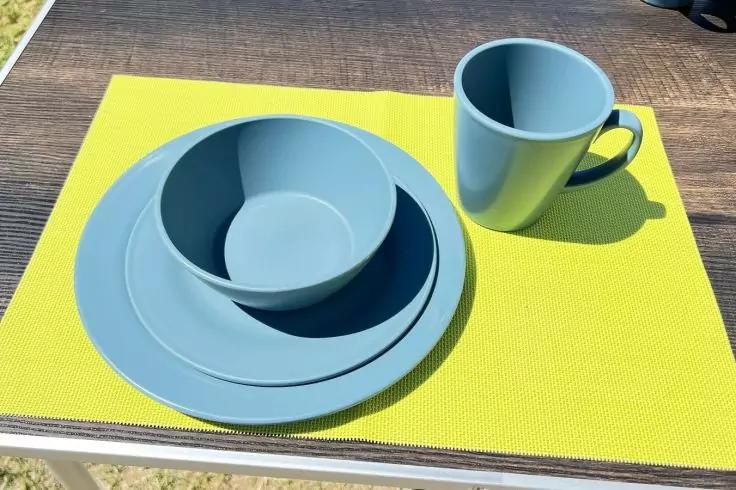 Hellblaue Teller und Schale gestapelt, dazu eine Tasse auf einem gelben Platzset auf einem Holztisch