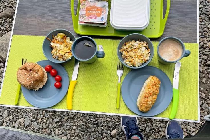 Zwei gedeckte Pl&auml;tze auf gr&uuml;nen Tischsets: blaue Teller mit einem Sesambr&ouml;tchen und einem l&auml;nglichen Geb&auml;ck, zwei Schalen mit gelbem, kr&uuml;meligem Inhalt, zwei Tassen mit dunkler Fl&uuml;ssigkeit, Besteck mit gelben und gr&uuml;nen Griffen, wei&szlig;e Plastikdose mit