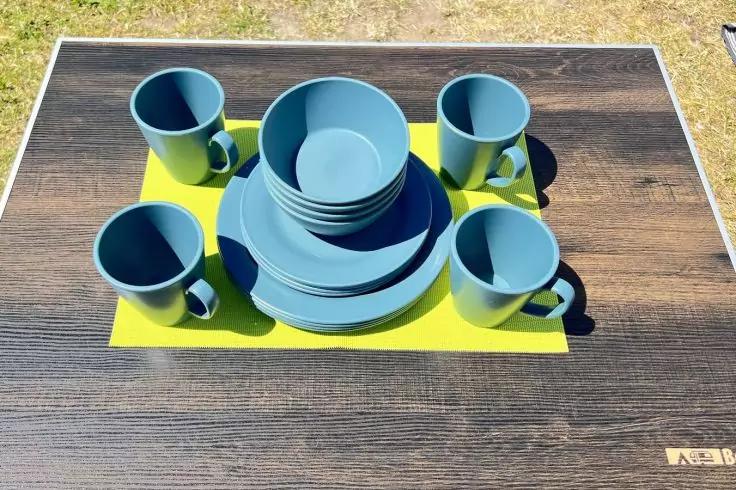 Dunkler Holztisch mit hellgr&uuml;nem rechteckigem Platzset. Auf dem Set gestapelte blaue Teller und Schalen in der Mitte und vier blaue Tassen an den Ecken. Gras im Hintergrund